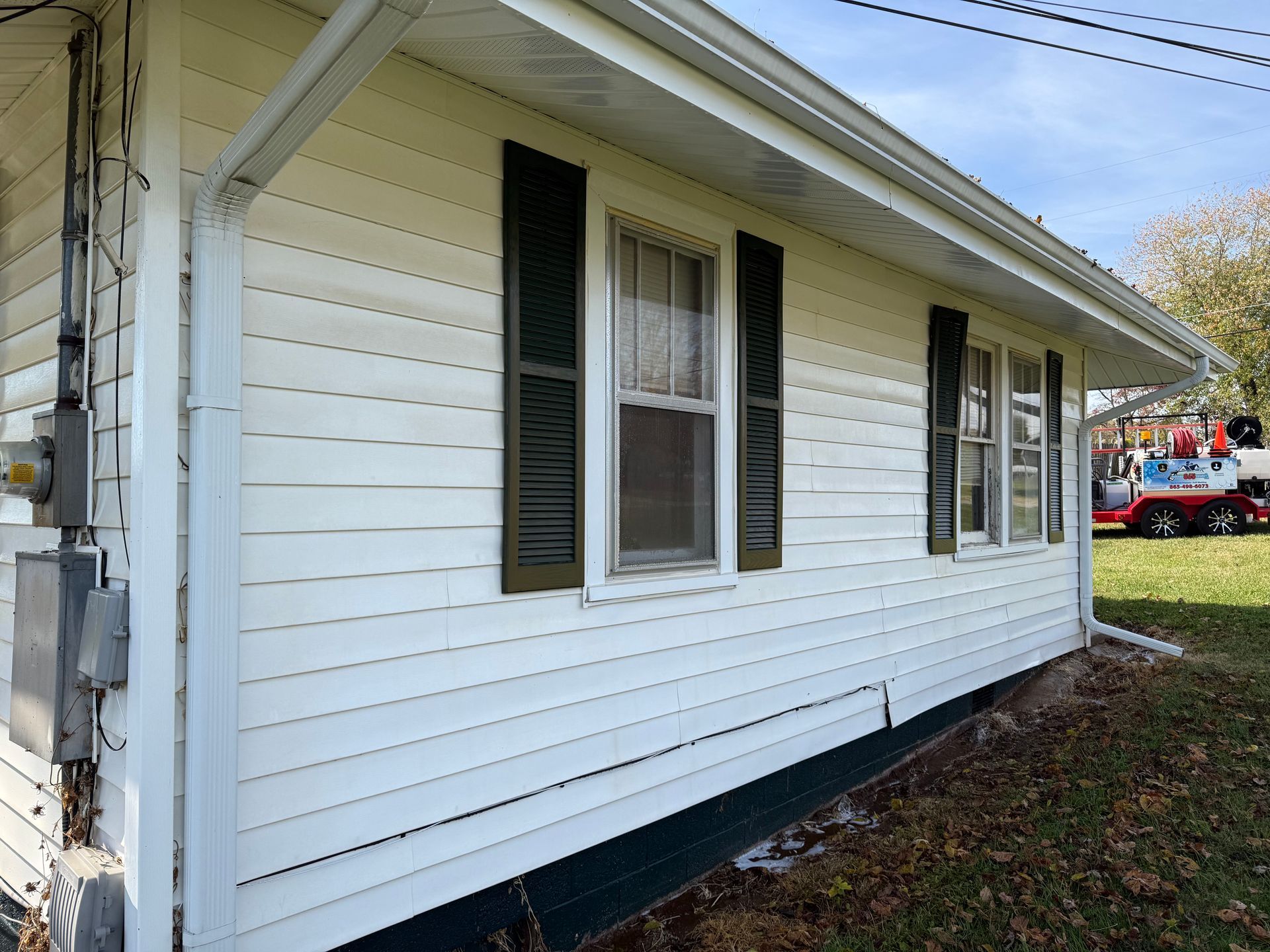 White-sided house with dark shutters, windows, and gutter. Green yard with a truck in the distance.