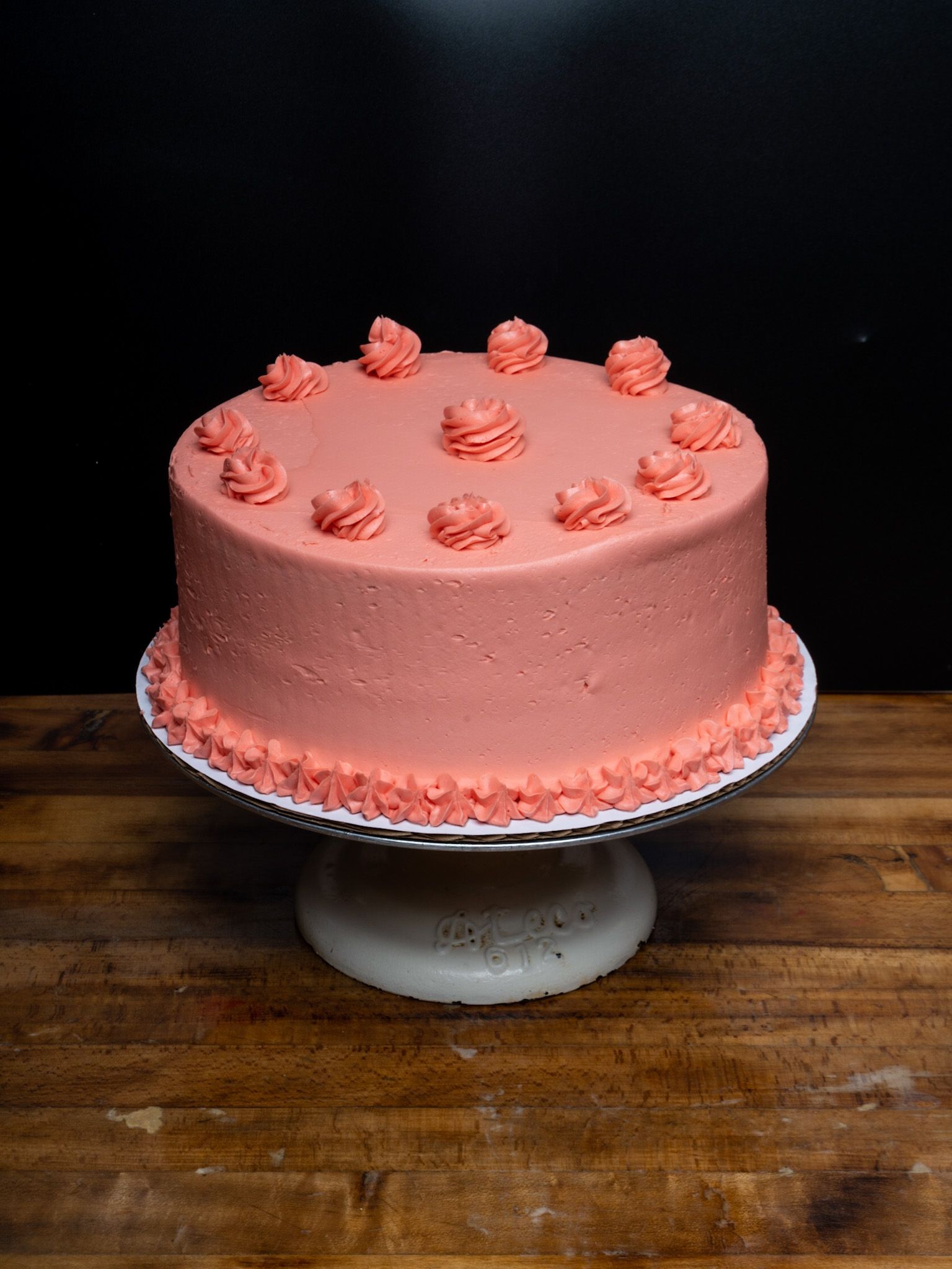 A pink cake is sitting on top of a white cake stand on a wooden table.