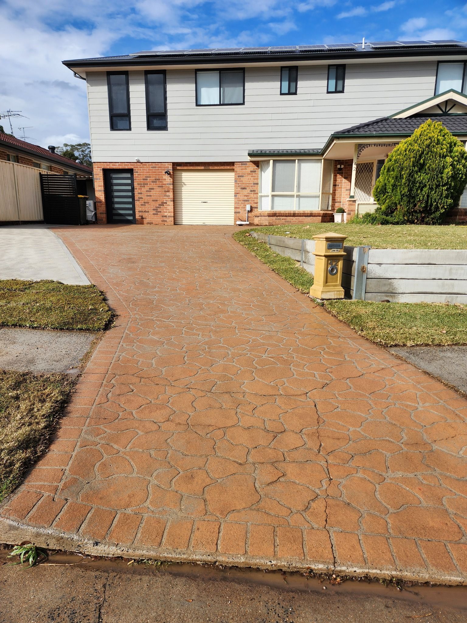 A suburban house with a patterned, reddish-brown concrete driveway leading to a garage under a cloudy blue sky.