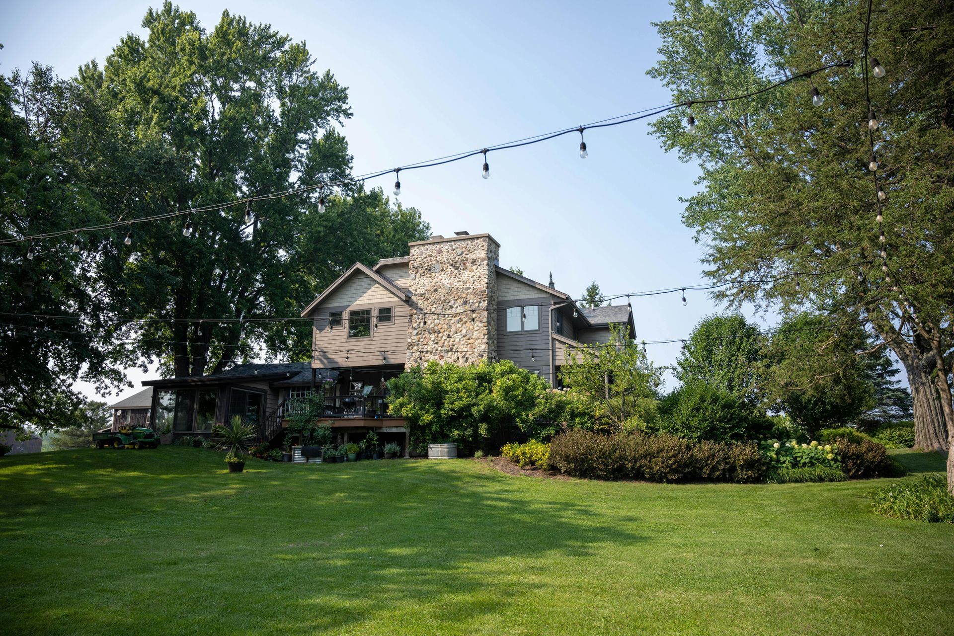A multi-story home with a stone chimney sits behind a green lawn, framed by large trees and strings of overhead lights.