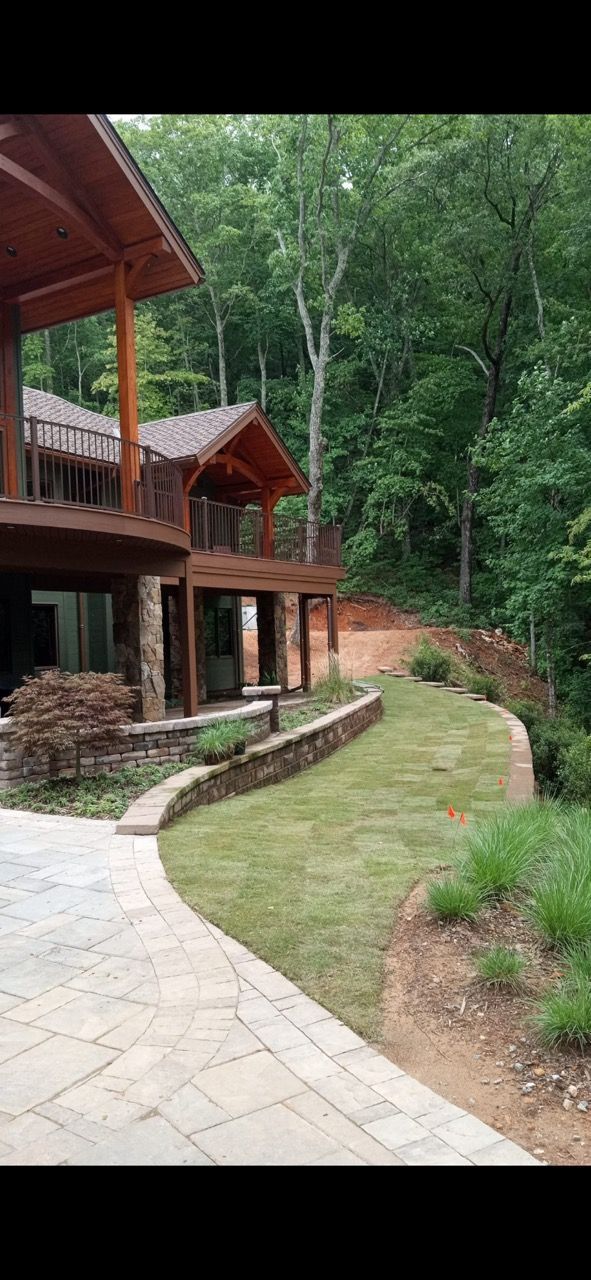 A multi-story wooden house with a stone foundation and patio sits beside a curved lawn and lush forest.