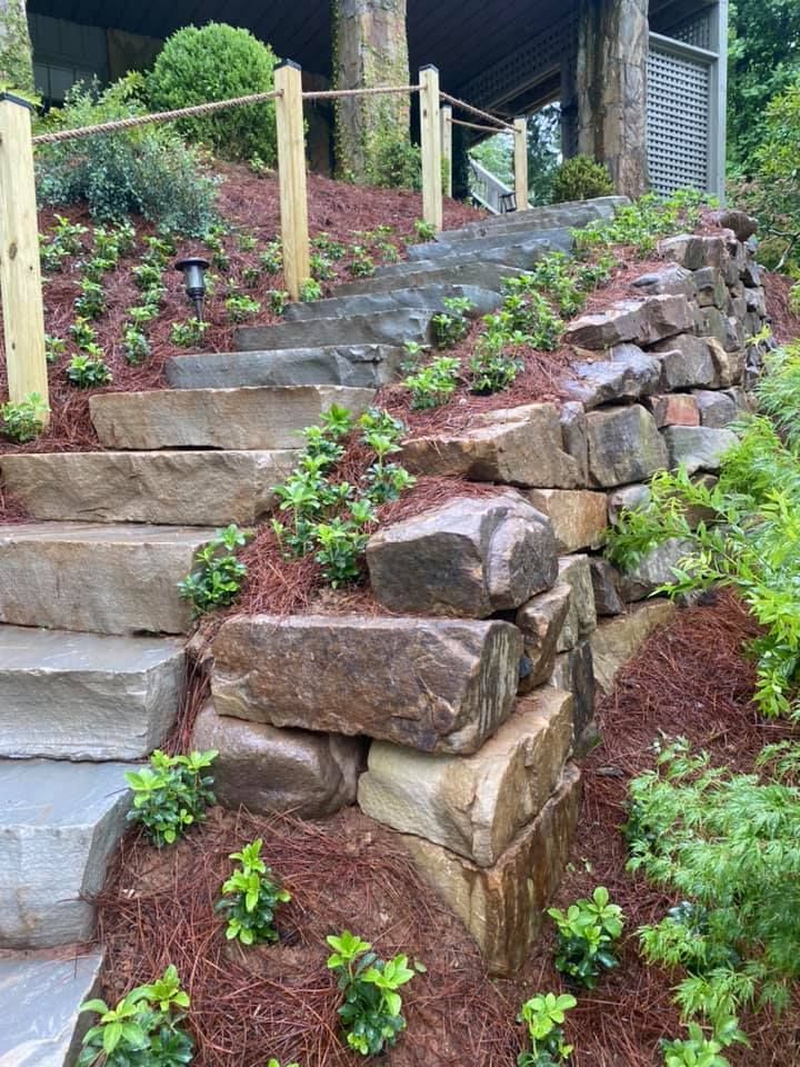 Stone steps and a terraced garden slope with rock retaining walls and green plants alongside a house