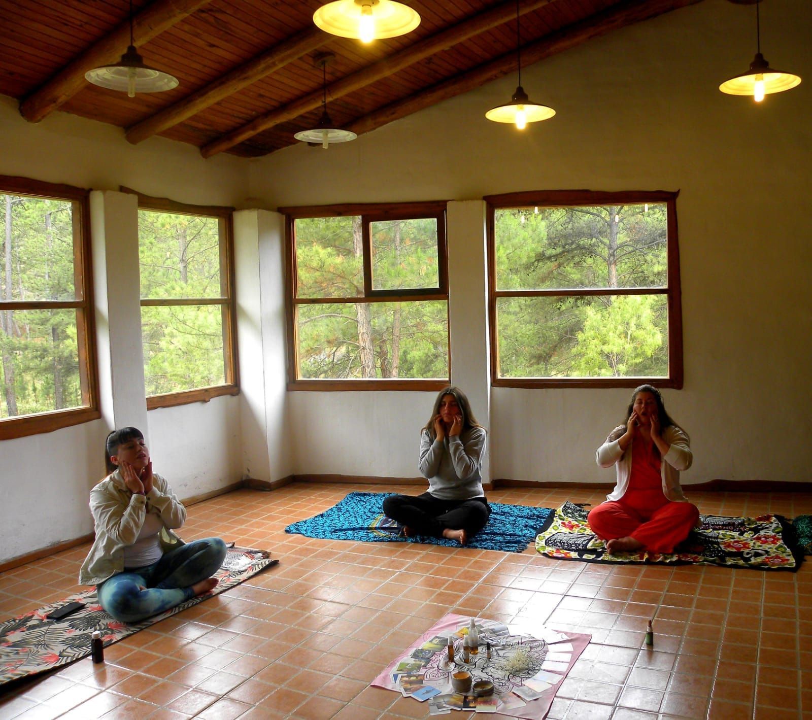 Tres personas sentadas sobre esteras, con las manos cerca de la cara, en una habitación con ventanas y luces colgantes.