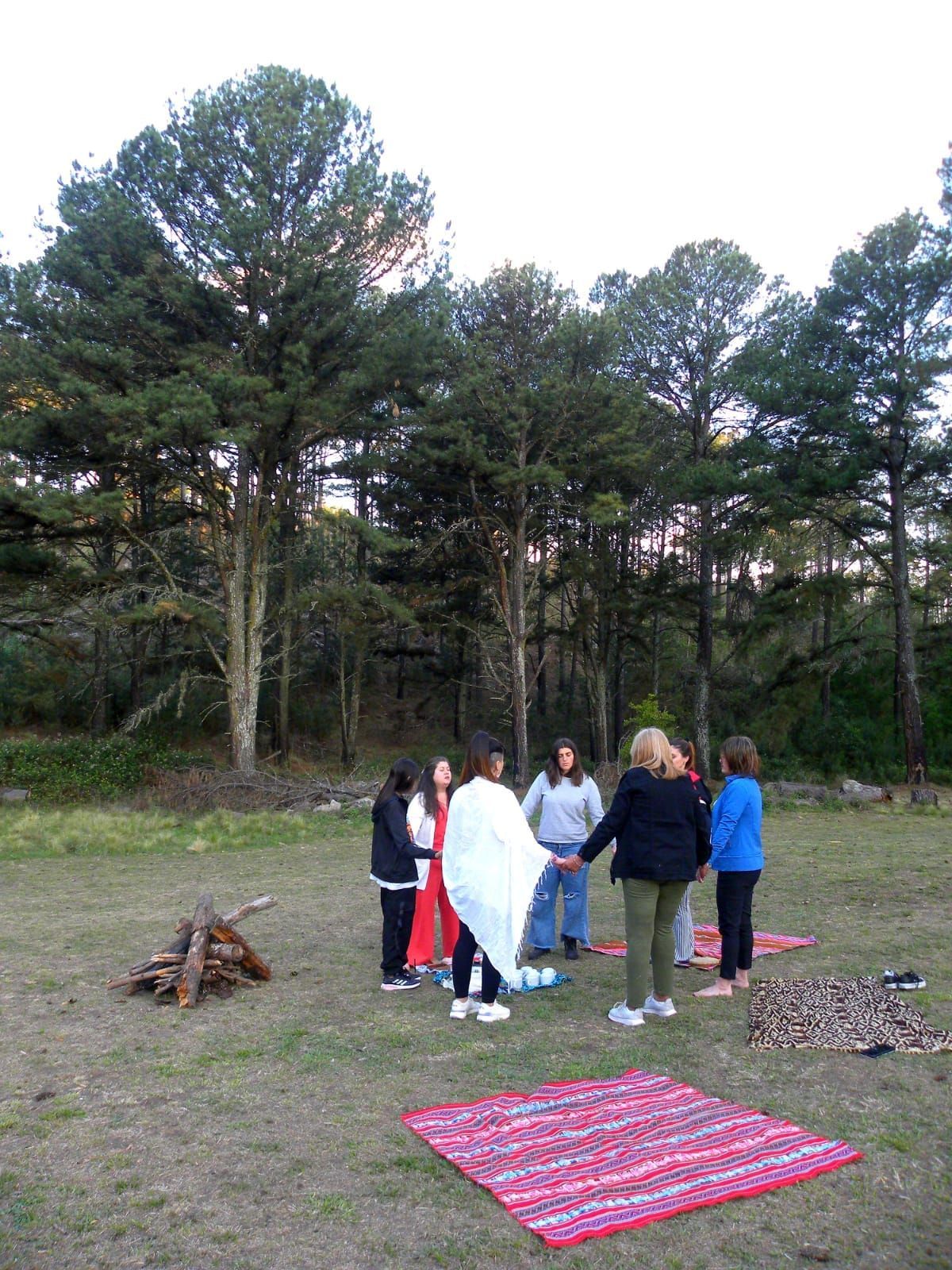 Personas tomadas de la mano formando un círculo alrededor de un fogón, en un claro cubierto de hierba y rodeado de árboles.