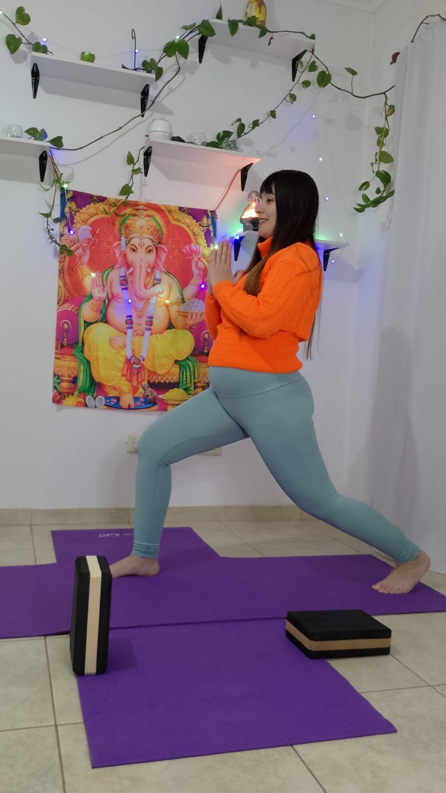 Mujer con top naranja y leggings azules en postura de yoga, manos juntas, habitación con colchonetas.