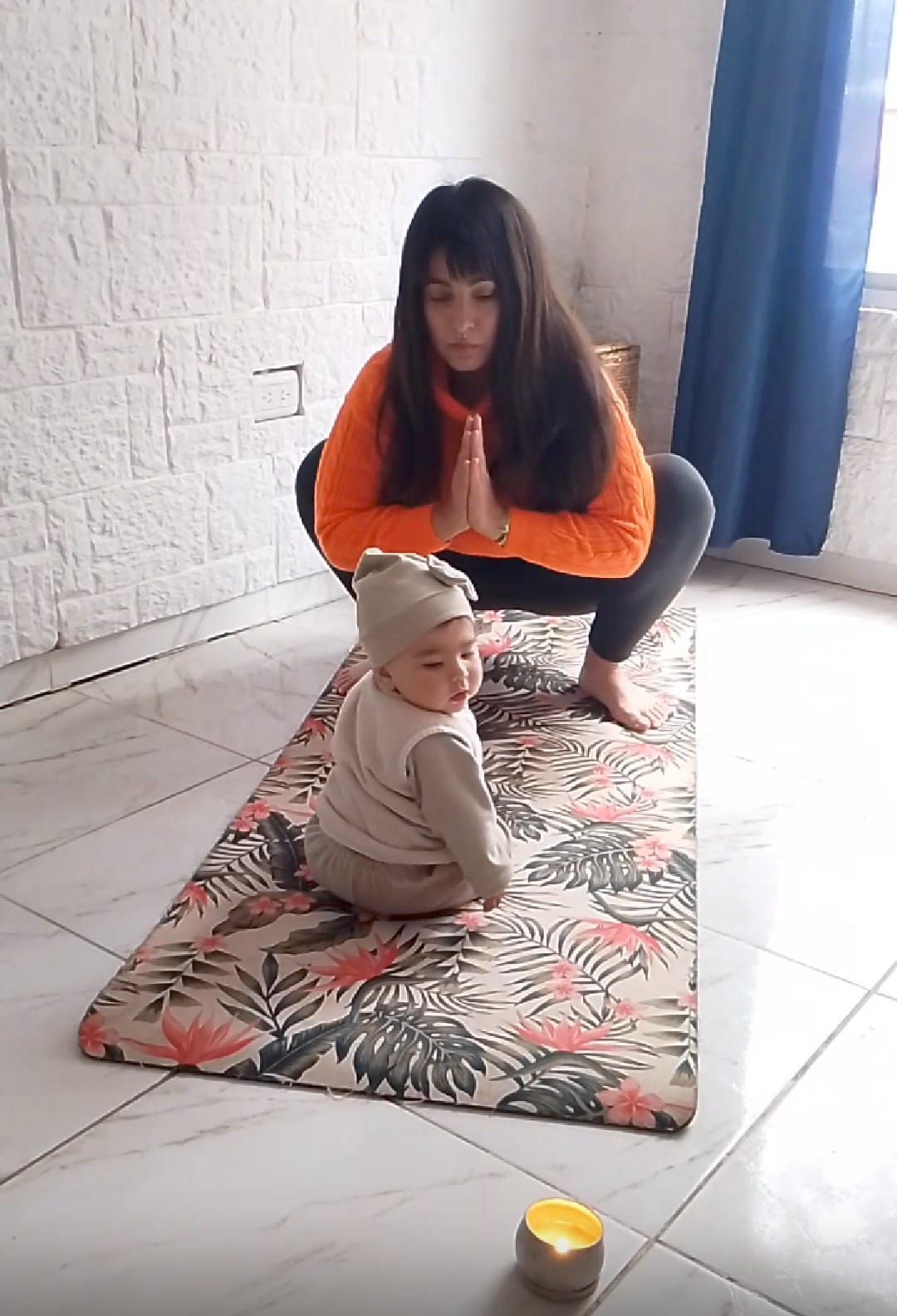 Mujer con top naranja haciendo yoga, niño con gorro sentado en la esterilla de yoga.
