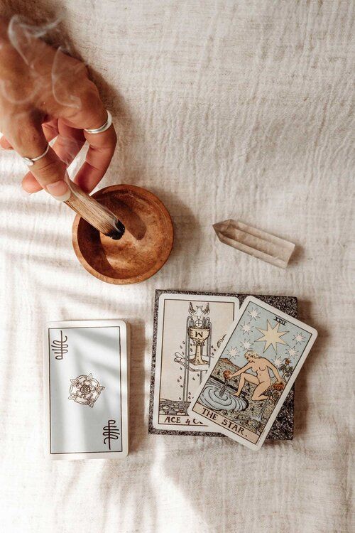 Mano sosteniendo palo santo ardiendo, cartas del tarot, cristal y cuenco sobre un paño blanco.