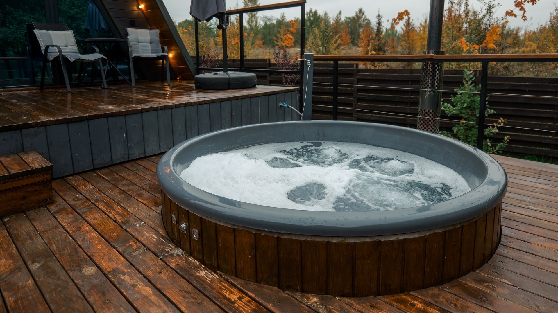 A circular gray hot tub filled with bubbling water on a wooden deck next to an A-frame cabin in a forested setting.