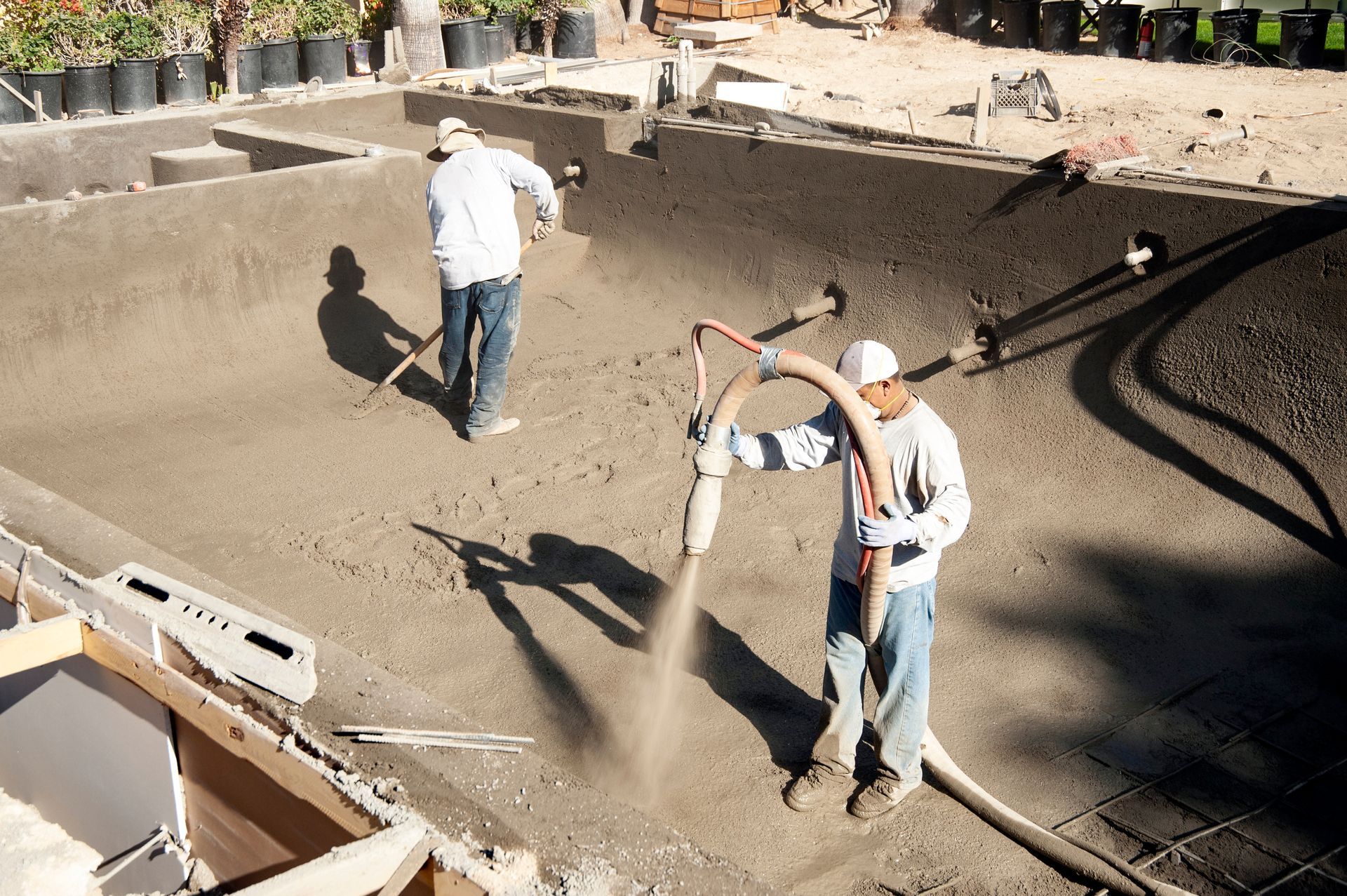 Two workers use a hose to spray concrete into the hollowed-out shape of a residential swimming pool construction site.