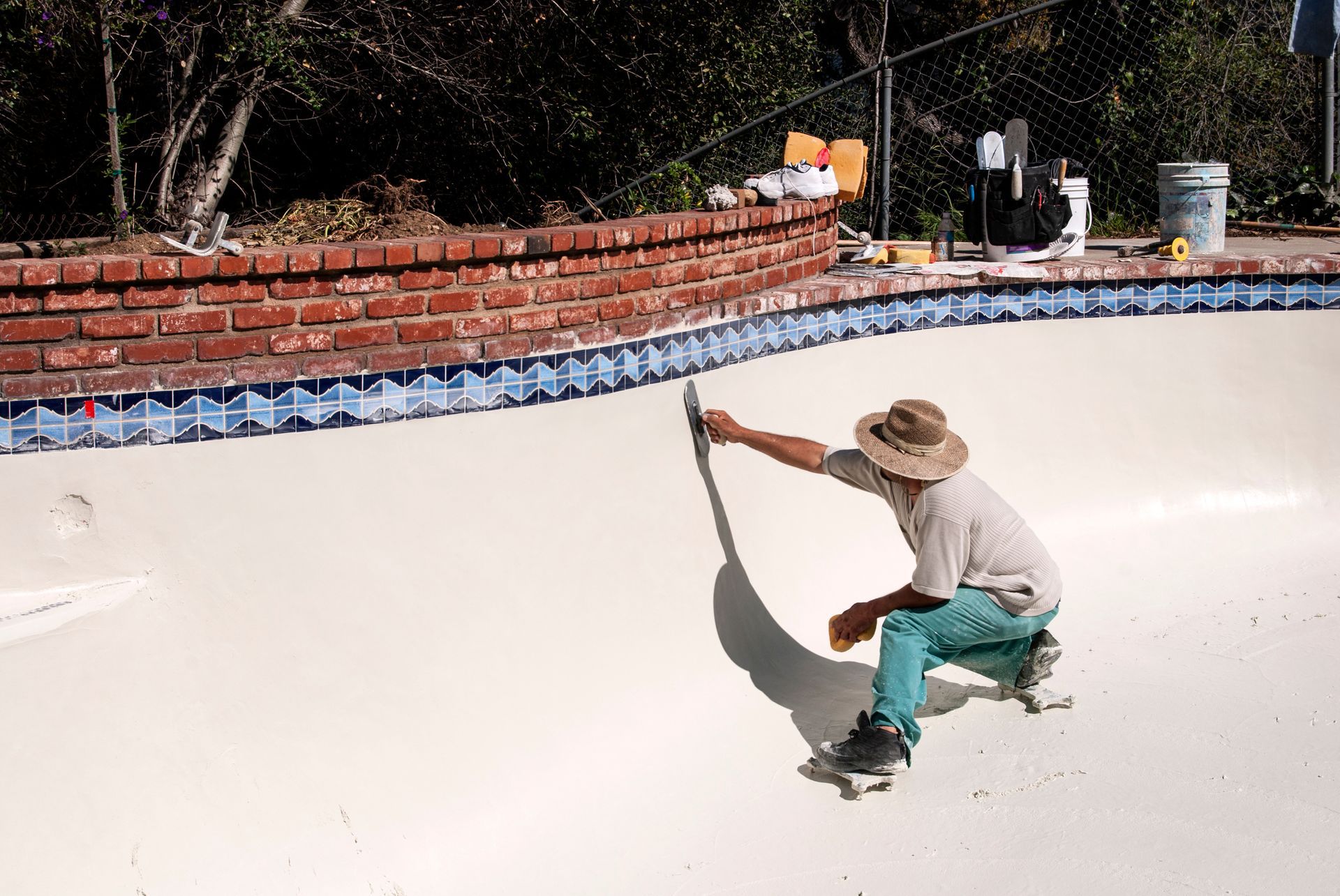 A worker in a hat and turquoise pants trowels white plaster onto the interior wall of an empty swimming pool.