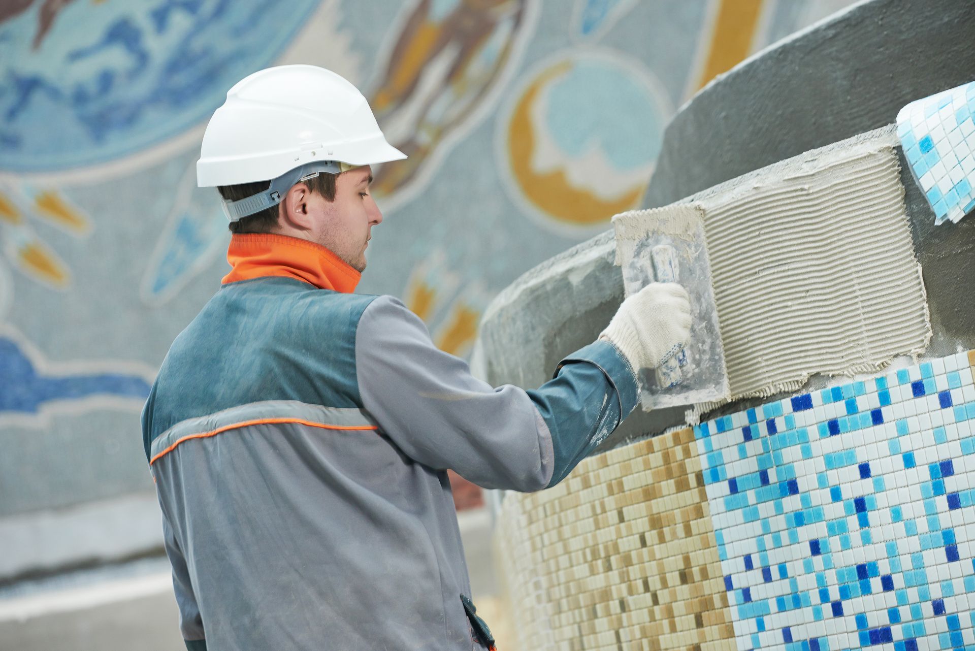 A construction worker in a white hard hat applying adhesive with a trowel to a tiled wall.
