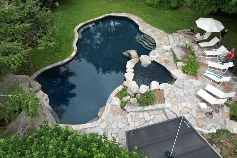 An aerial view of a natural-shaped swimming pool with stone coping and a hot tub, surrounded by a patio and lounge chairs.