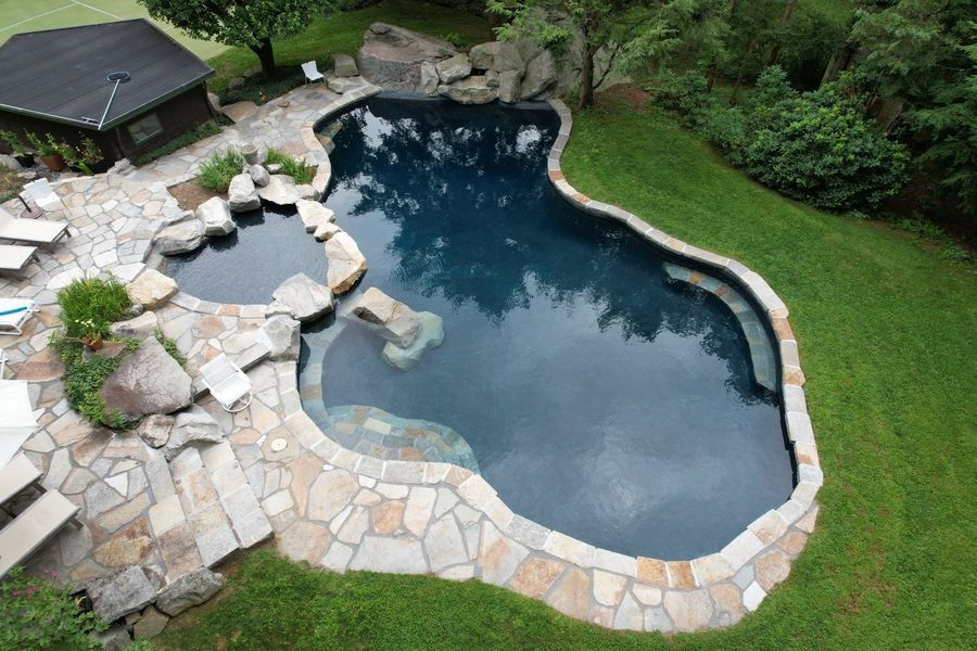 A high-angle view of a dark, free-form swimming pool with a stone patio, adjacent hot tub, and lush green lawn.
