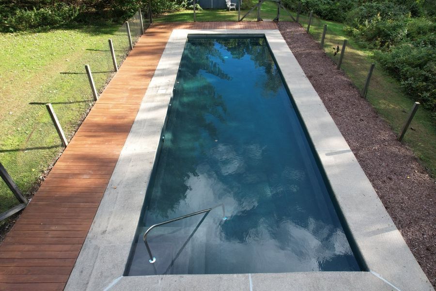 A narrow rectangular swimming pool with dark blue water, flanked by a wooden deck on one side and gravel on the other.