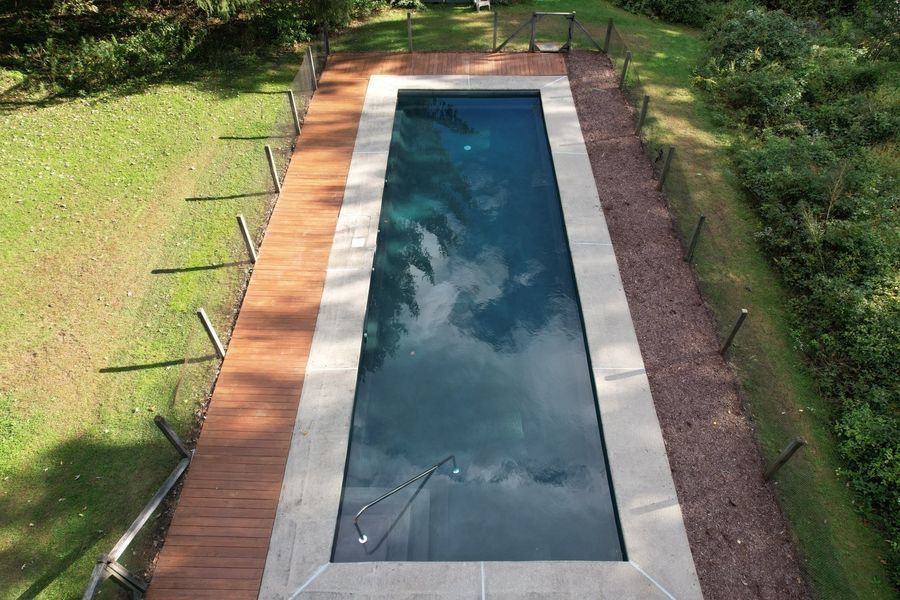 A rectangular swimming pool with dark water and stone coping, bordered by wooden decking and gravel in a garden setting.