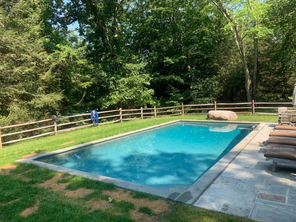 A rectangular swimming pool with blue water sits in a green lawn next to a patio and a wooden fence near a forest.