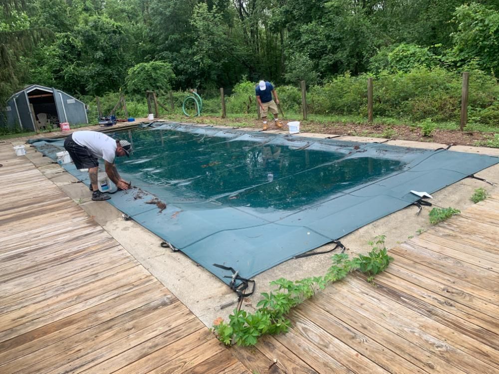 Two people work on securing a large dark green safety cover over a backyard swimming pool surrounded by a wooden deck.