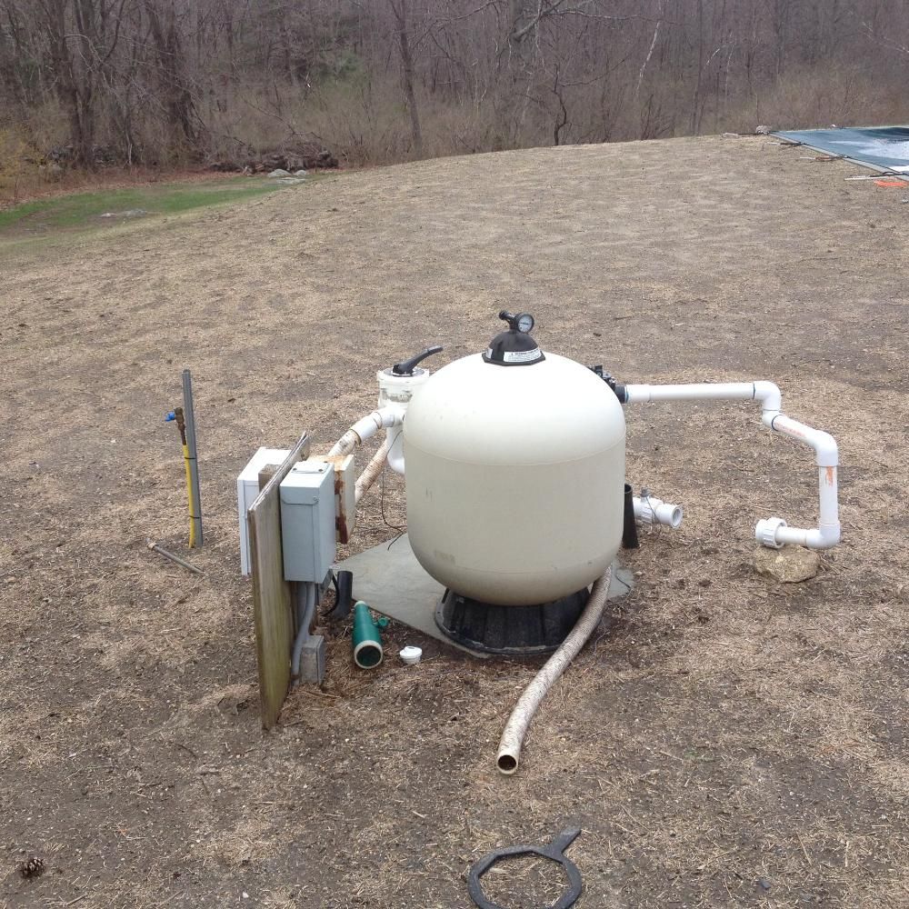 A white pool sand filter system with connected pipes and electrical box sits outdoors on a dirt patch near a pool cover.
