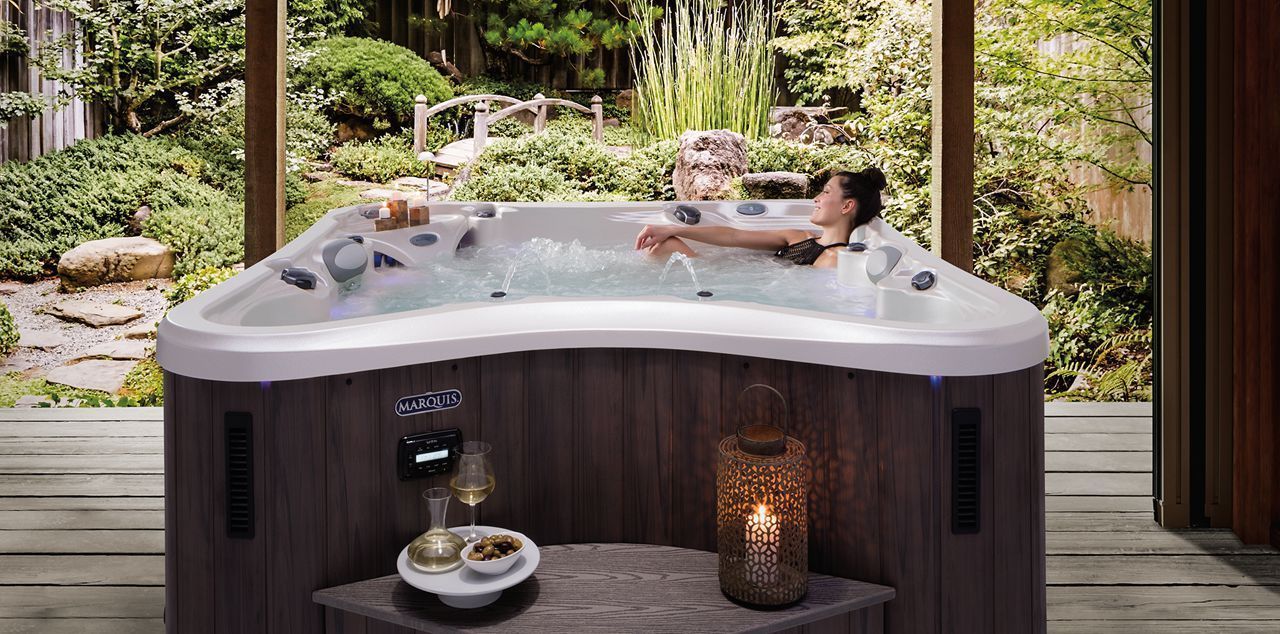 A person relaxes in a corner-shaped hot tub on a wooden deck overlooking a lush garden.