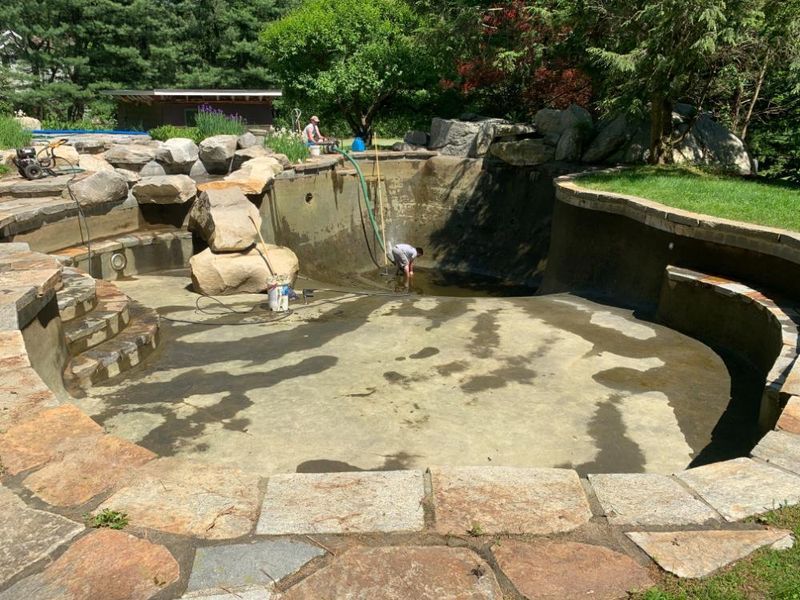 A worker cleans a drained stone-lined pond or swimming pool with a hose in a garden setting.