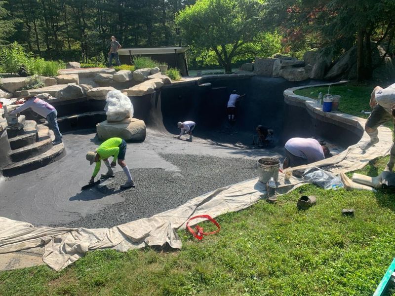 Workers construct a backyard pond, spreading gravel over the dark pool liner while surrounded by landscape stone.
