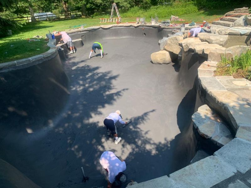 Four workers in a pool basin are applying a dark finish to the walls and floor, surrounded by stone landscaping.