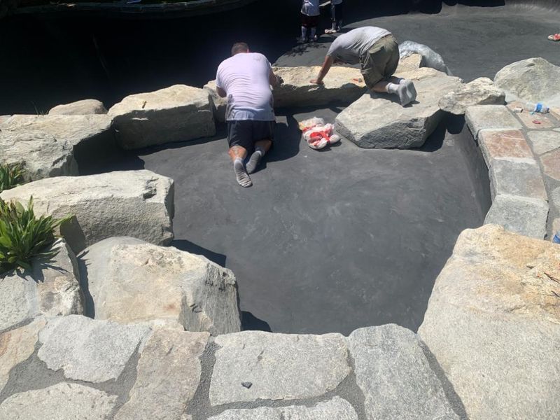 Two people kneel inside a stone-bordered pool, applying sealant to the dark gray interior surface.