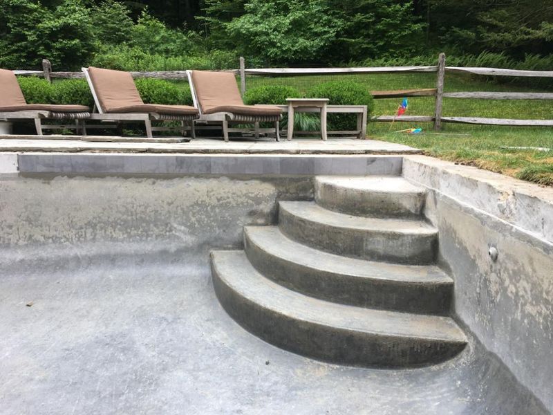 Empty swimming pool with curved concrete steps leading to the patio, where lounge chairs sit on the grass.