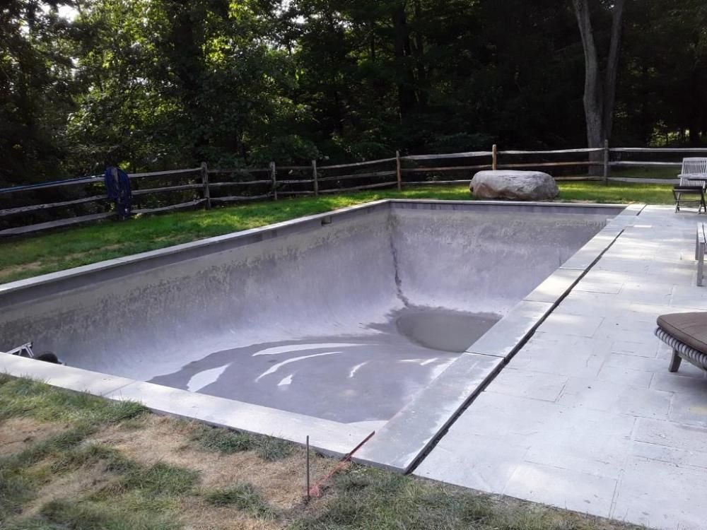 An empty concrete swimming pool in a grassy backyard with a split-rail fence and a large rock nearby.