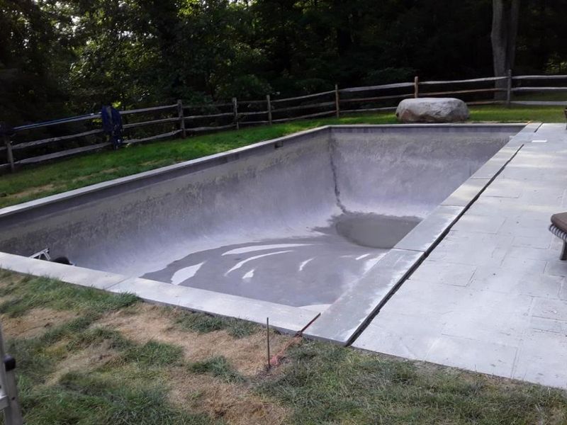 An empty, newly plastered rectangular swimming pool in a backyard with a wooden fence and a stone patio.