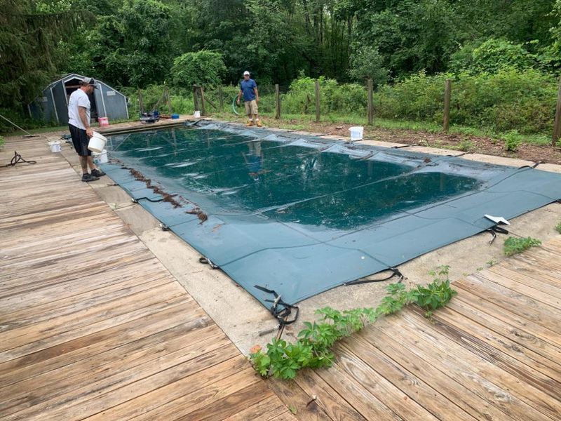 Two people working on a pool covered by a large, dark green tarp on a wooden deck in a grassy, wooded outdoor area.