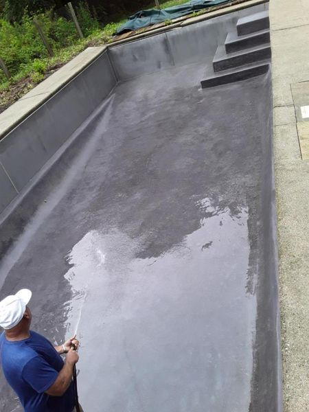 A person in a blue shirt sprays water into an empty, dark gray rectangular swimming pool with built-in stairs.