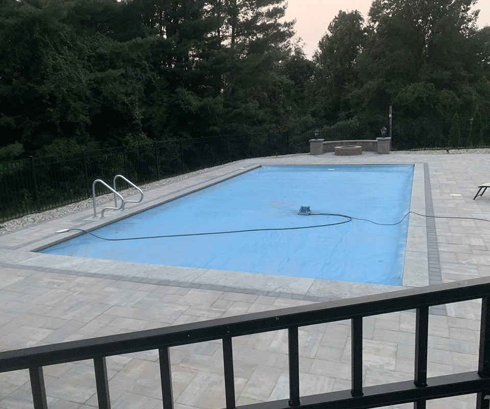 A rectangular swimming pool covered with a blue tarp, surrounded by grey stone pavers in a backyard setting.