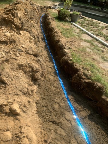 A bright blue utility warning tape runs along the center of a freshly dug dirt trench in a residential yard.