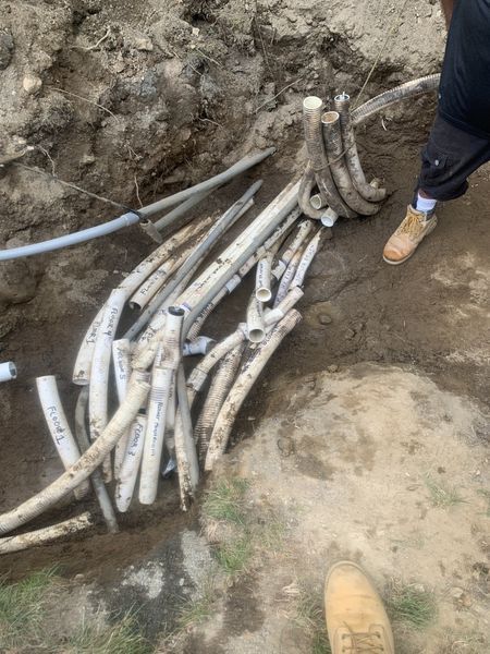 A person stands near a trench containing a complex cluster of white PVC pipes and various buried electrical conduits.
