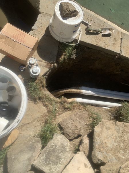 A deep hole in the ground reveals several white PVC pipes next to stone landscaping, a bucket, and supplies.