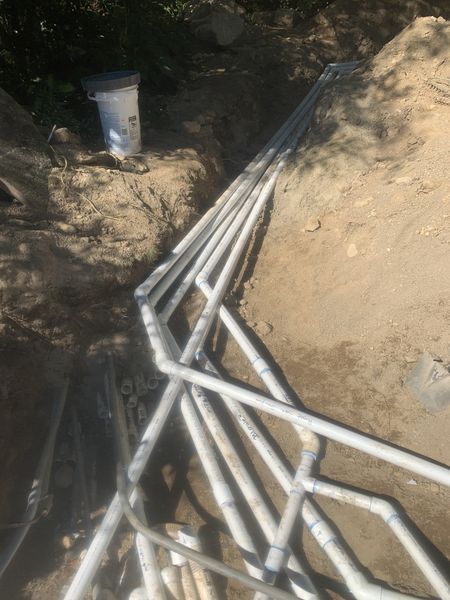 A trench in the dirt containing several white PVC pipes connected with elbows and couplings.