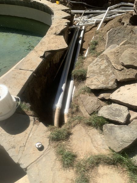 A trench dug beside a pool containing exposed white PVC plumbing pipes and stacked stones nearby.
