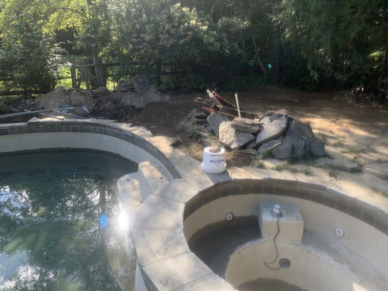 A half-finished outdoor swimming pool and spa area under construction, surrounded by piles of dirt, rock, and debris.