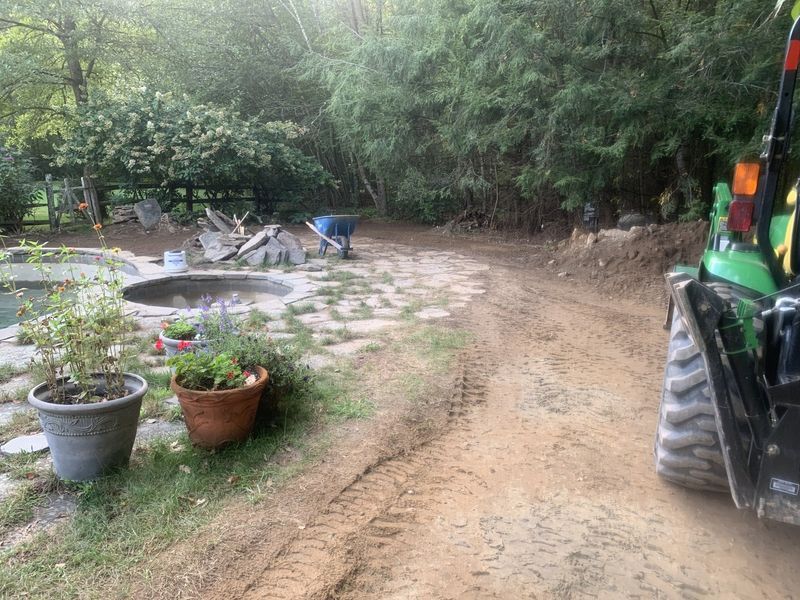 A person works on a stone patio near a pool, with a green tractor parked on a dirt path in the foreground.