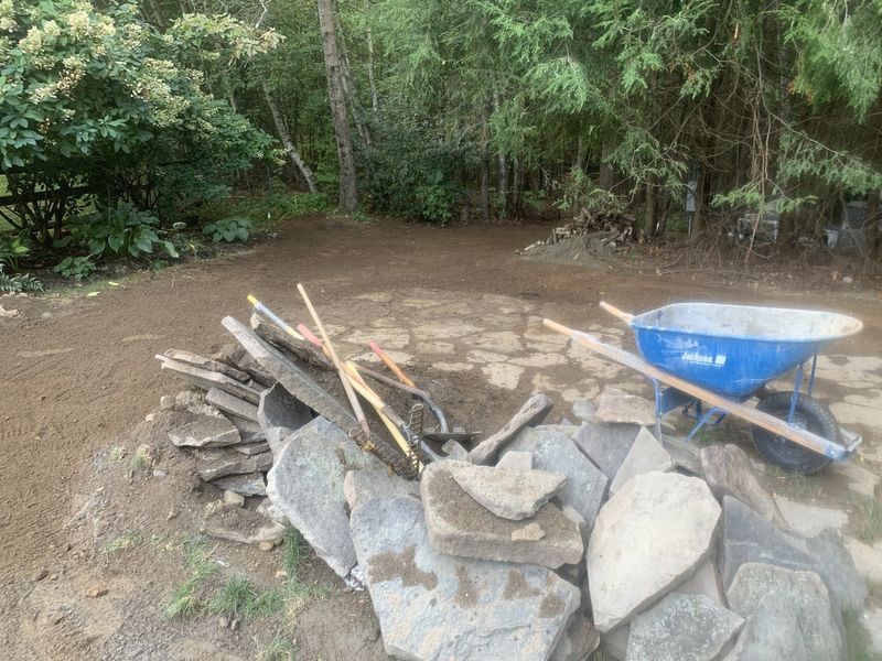 A pile of flat, grey stones for landscaping sits on dirt next to a blue wheelbarrow in a wooded area.