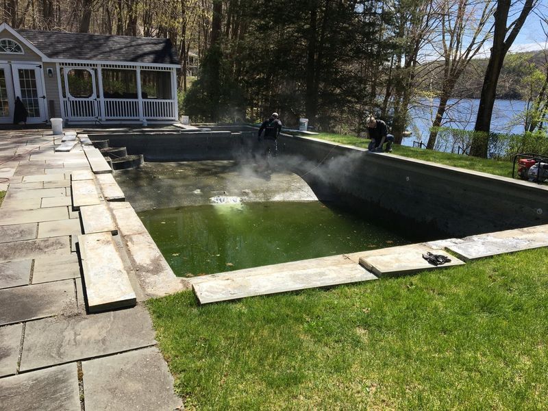 Two workers pressure-wash the stained concrete interior of a drained backyard swimming pool.