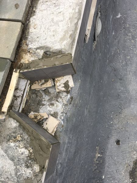 A section of wall showing loose, detached gray tiles, exposed cement, and debris on a dark gray foundation.