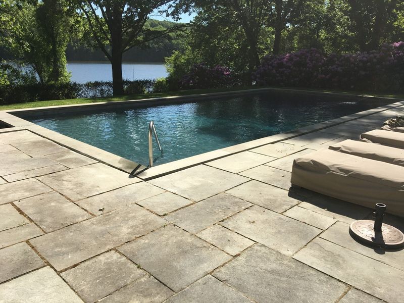 A rectangular swimming pool surrounded by stone patio tiles, featuring lounge chairs overlooking a lake and trees.