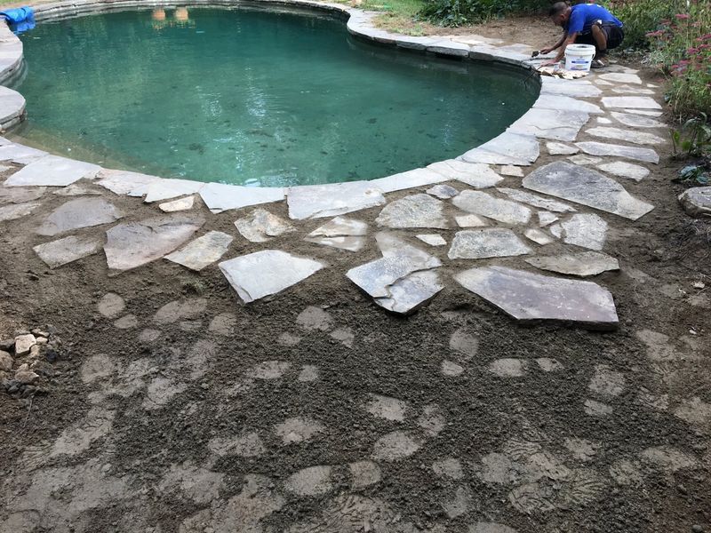 A person works on stone pavers surrounding a backyard swimming pool under construction.