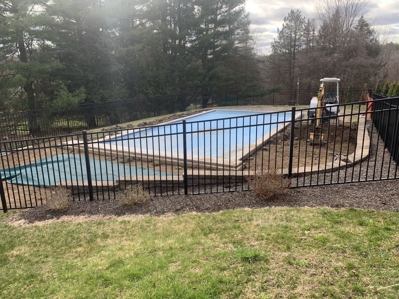 A blue rectangular swimming pool surrounded by a black metal fence, with a small excavator parked on the side.