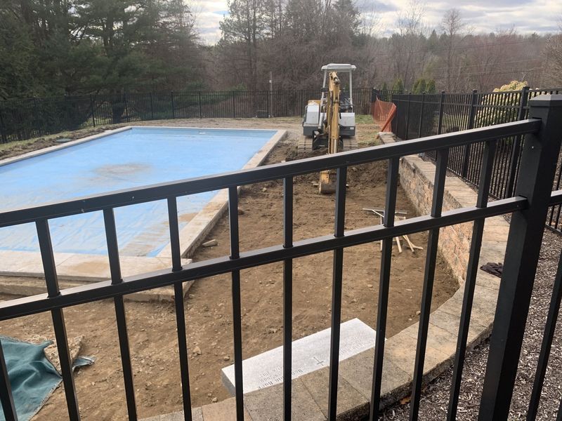 A construction excavator works in a dirt area next to a swimming pool with a blue cover, viewed through a black fence.