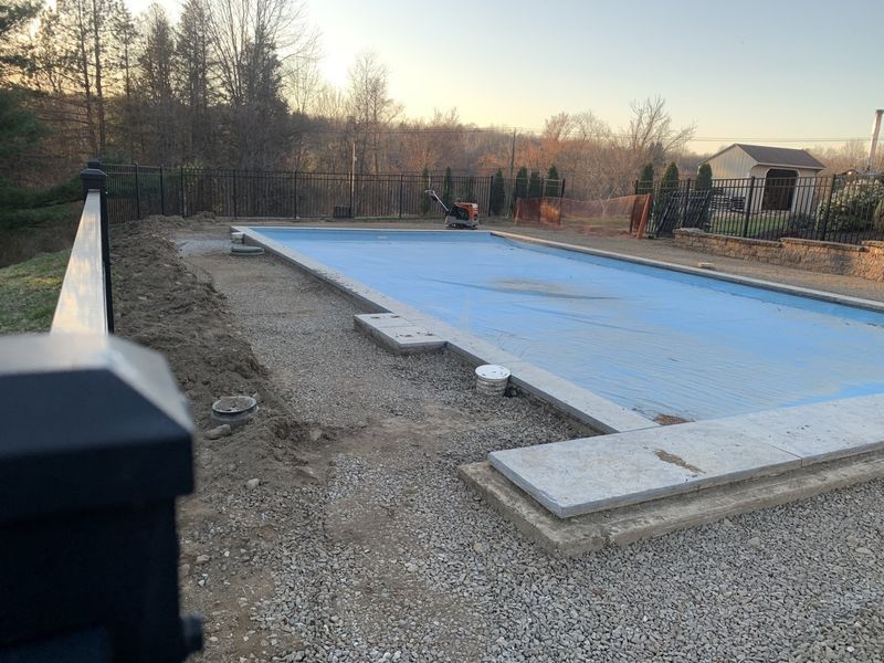 A rectangular backyard pool covered with a blue tarp, surrounded by gravel and stone coping under a clear sunset sky.