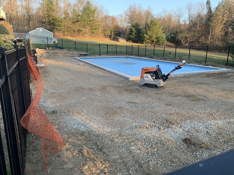 An orange plate compactor sits on gravel next to a rectangular swimming pool under a blue cover in a backyard.
