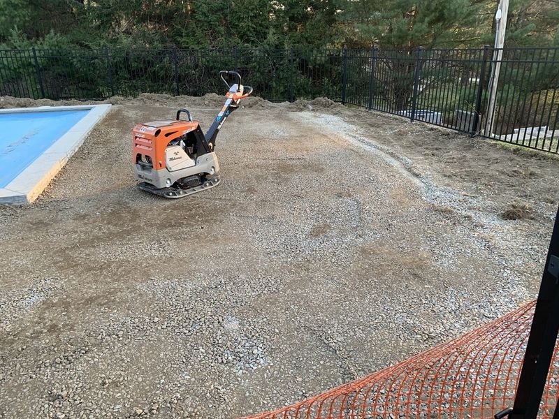 An orange compactor sits on a gravel patch beside a swimming pool, preparing the ground for construction.